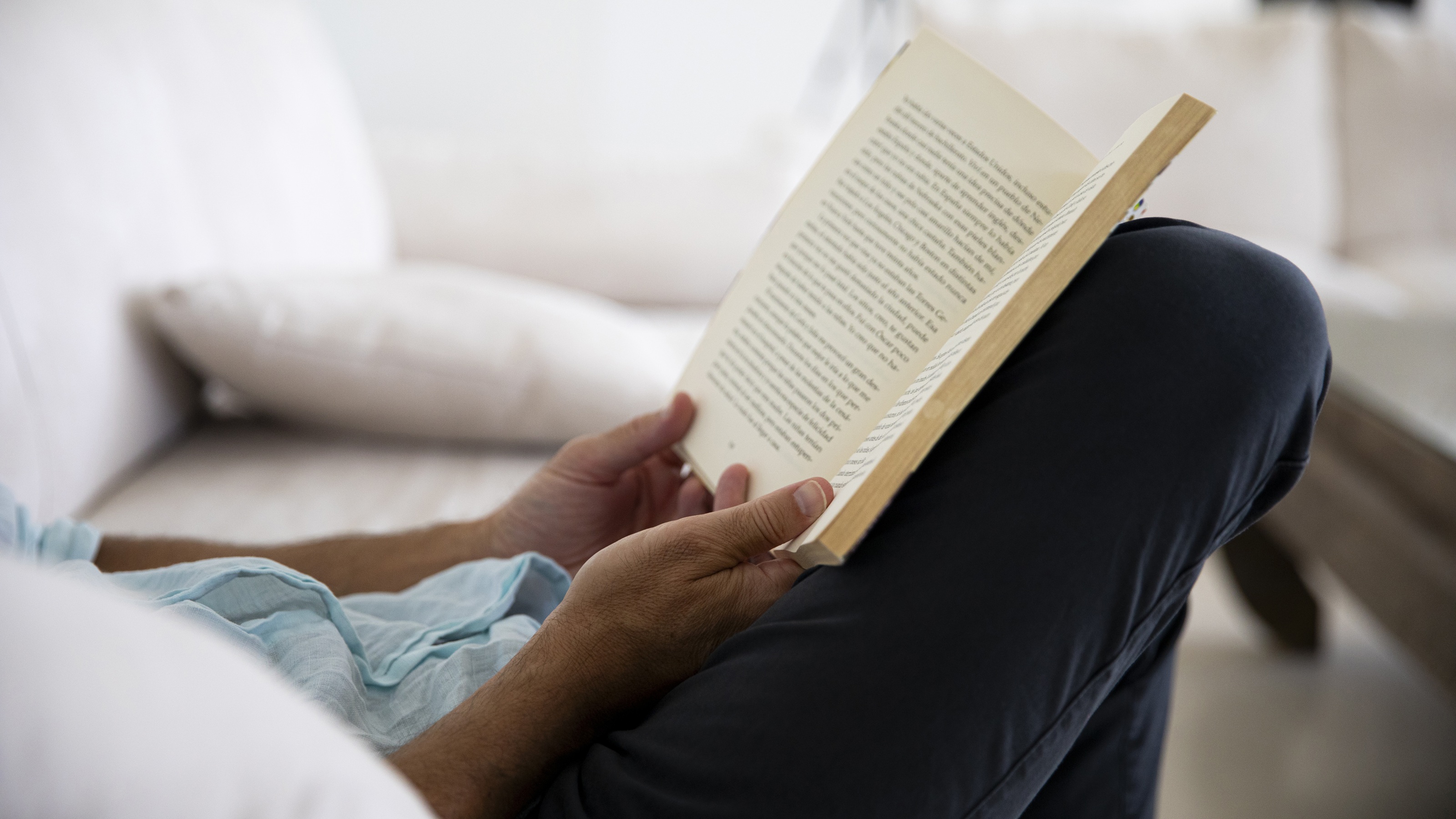 A man sits on a sofa in a quiet environment reading a book.