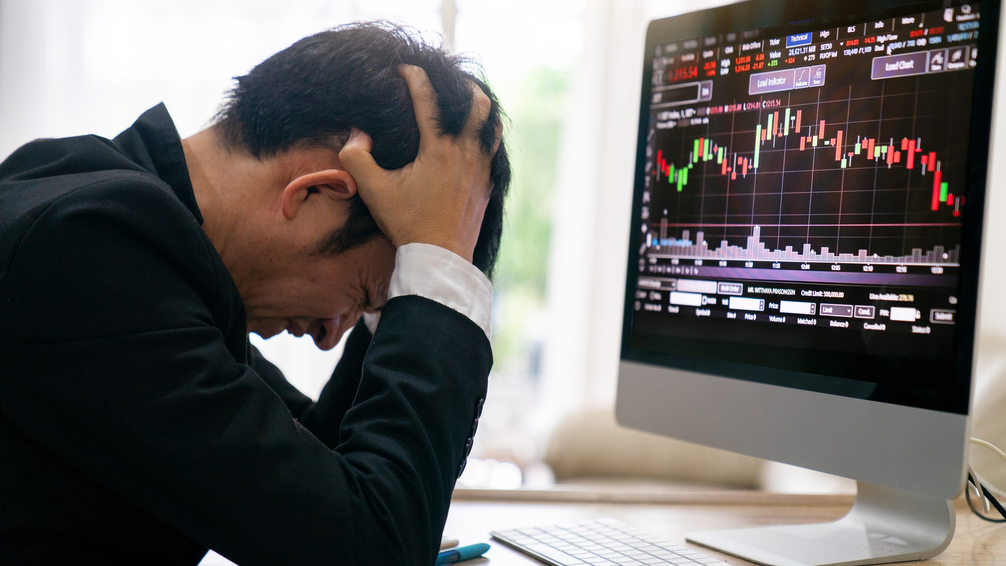 A frustrated investor sitting with his head in his hands in front of a computer screen showing a complex trading graph.