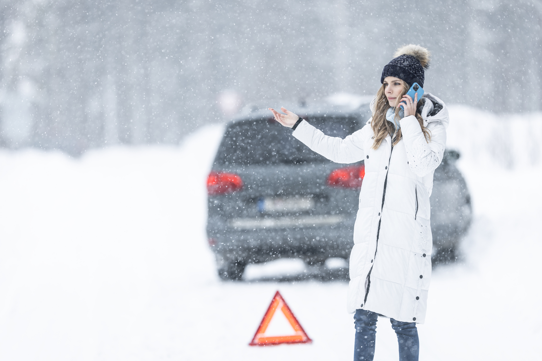 A driver standing by a car that has skidded off a snowy road, placing an emergency triangle and calling for help.