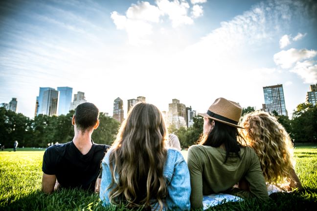 A group of friends sitting together on the grass in a city park with buildings in the background.