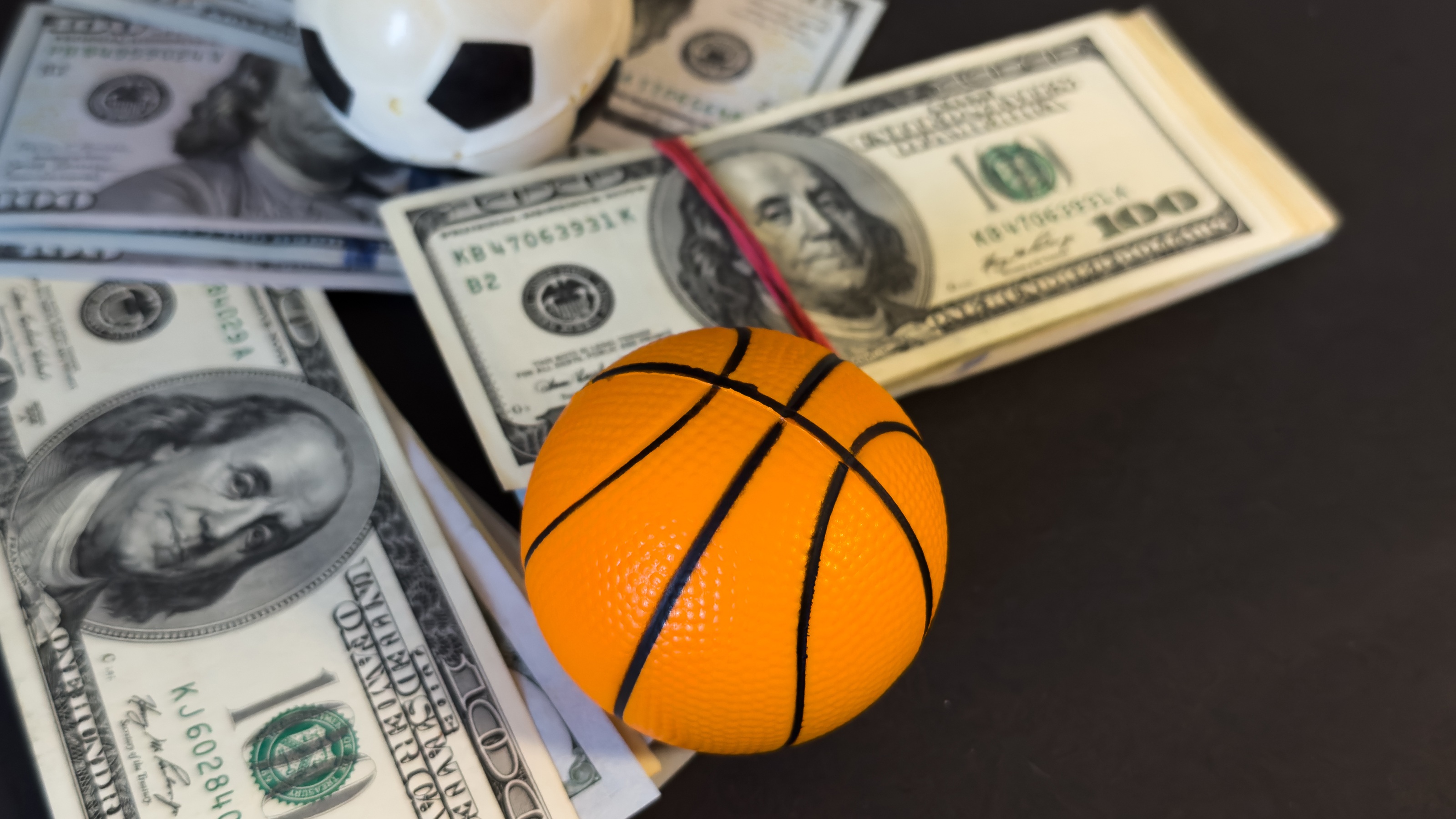 Stacks of cash placed under a soccer ball and a basketball on a flat surface.