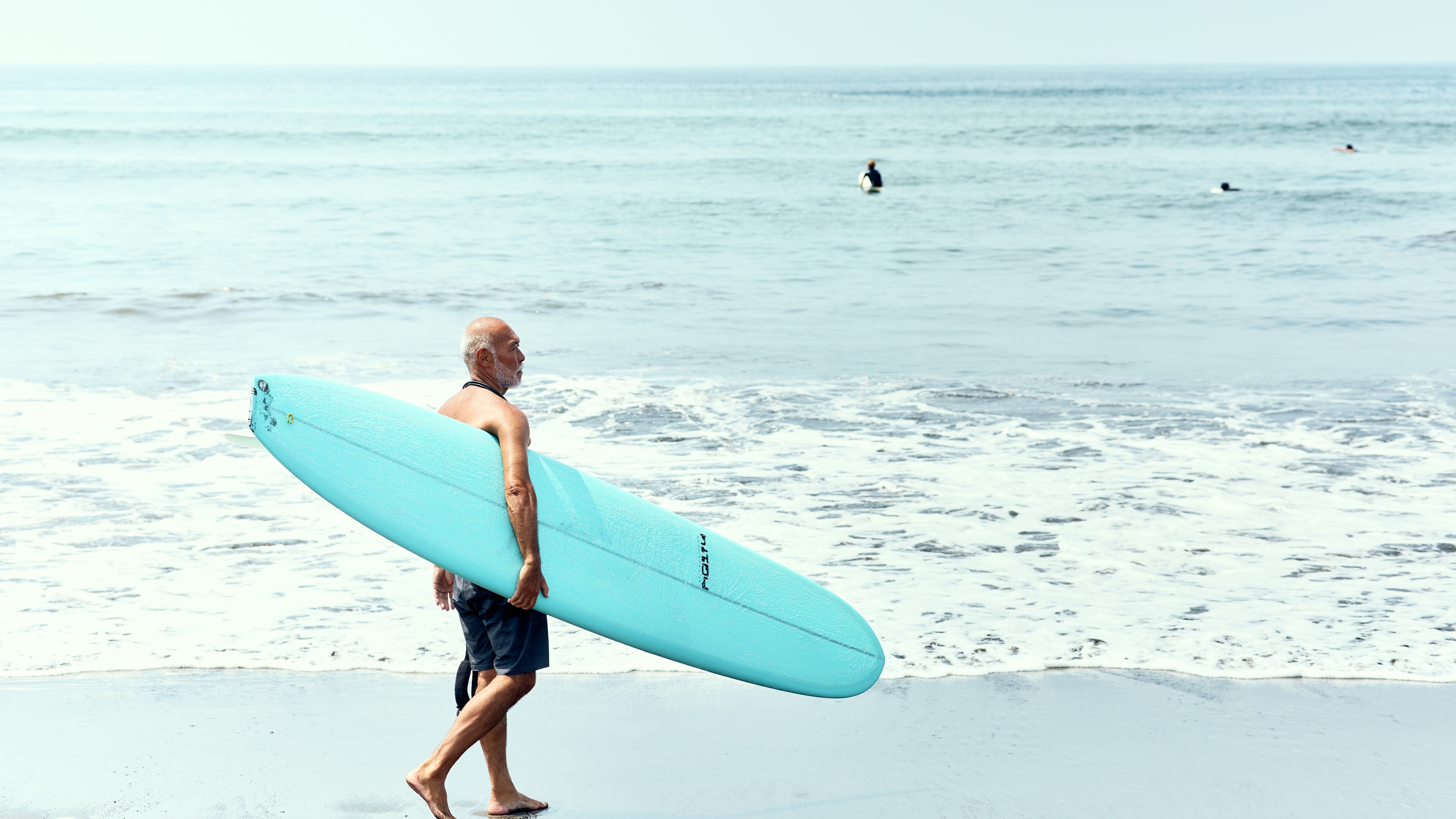 An active senior man carrying a surfboard on a beach during a bright day.